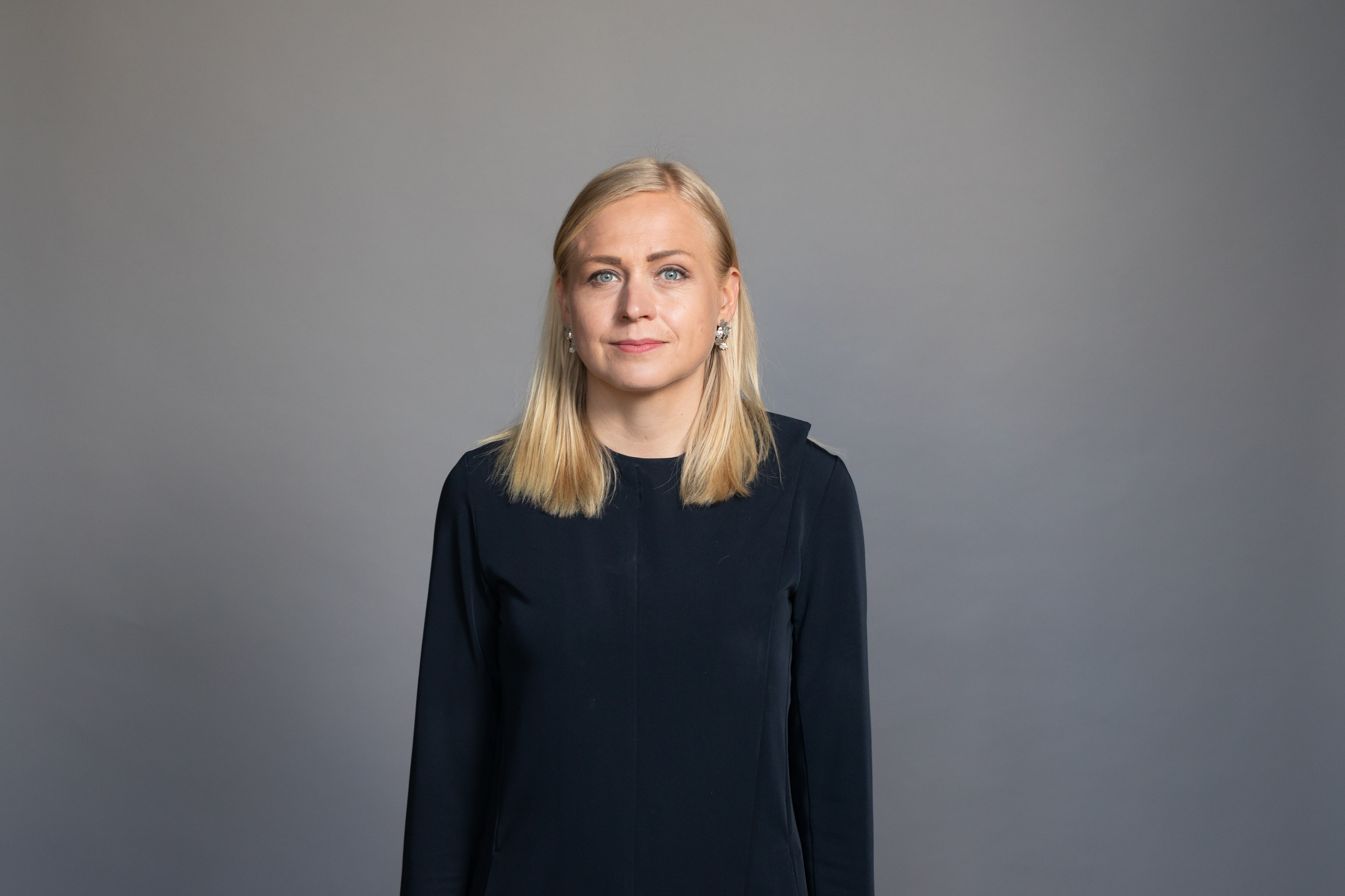 Arkistokuva: Vaaleahiuksinen nainen, jolla on tummansininen pitkähihainen paita ja korvakorut, katsoo suoraan kameraan harmaata taustaa vasten.