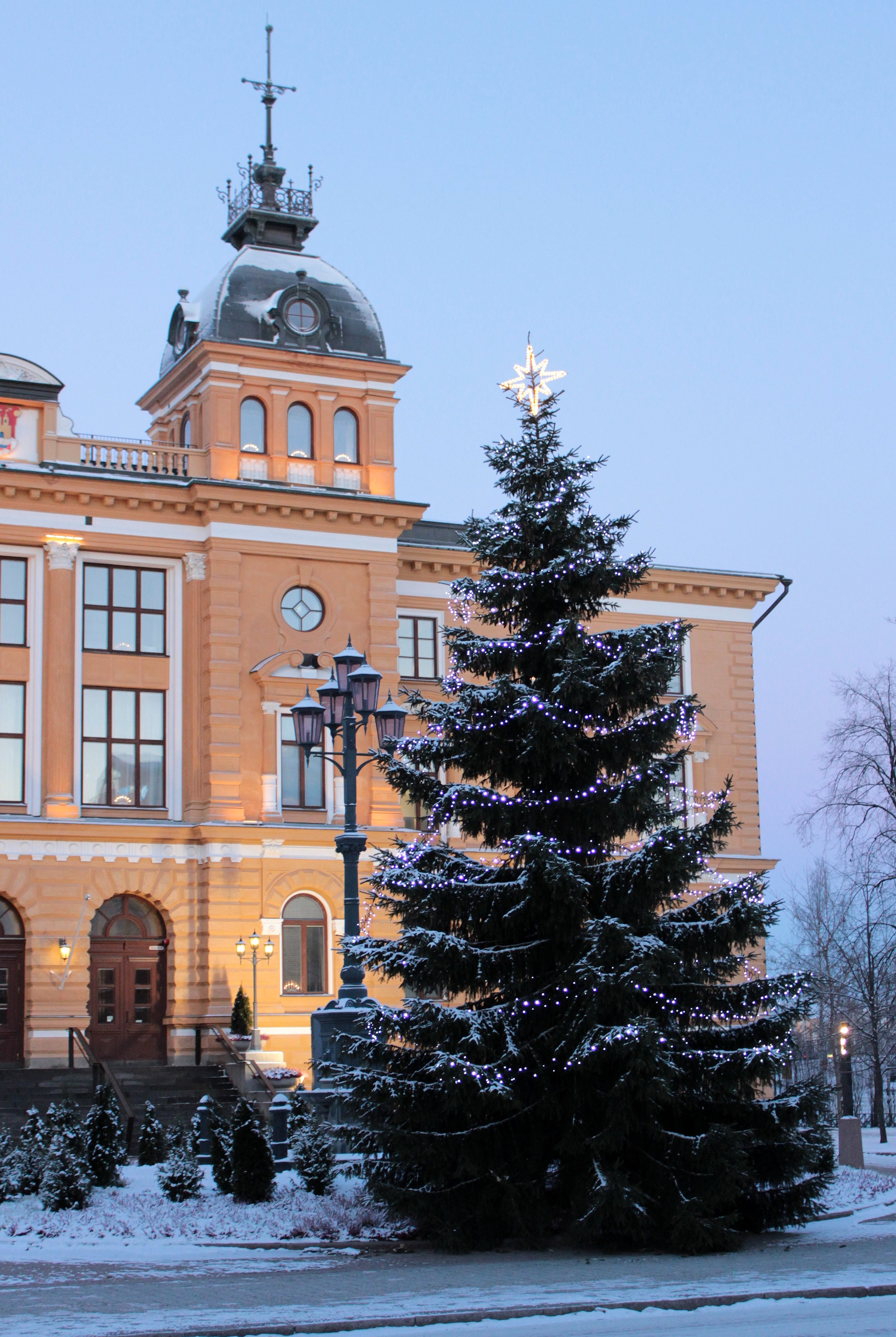 Arkistokuva: Suuri, lumipeitteinen joulukuusi valkoisine valoineen ja tähden kanssa seisoo vaalean oranssin rakennuksen edessä.