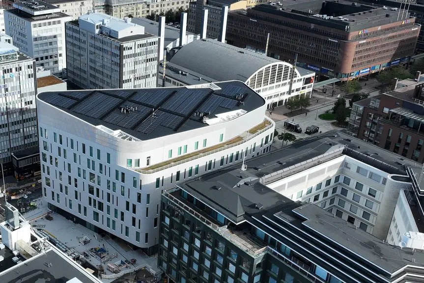 Arkistokuva: Valkoinen monikerroksinen rakennus aurinkopaneeleineen näkyy ilmasta muiden rakennusten ja katualueiden keskellä.