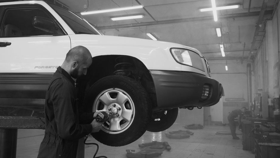 Arkistokuva: Mekaanikko työskentelee auton pyörän parissa korjaamolla. Auto on nosturilla, ja mekaanikko käyttää työkalua pyörään.