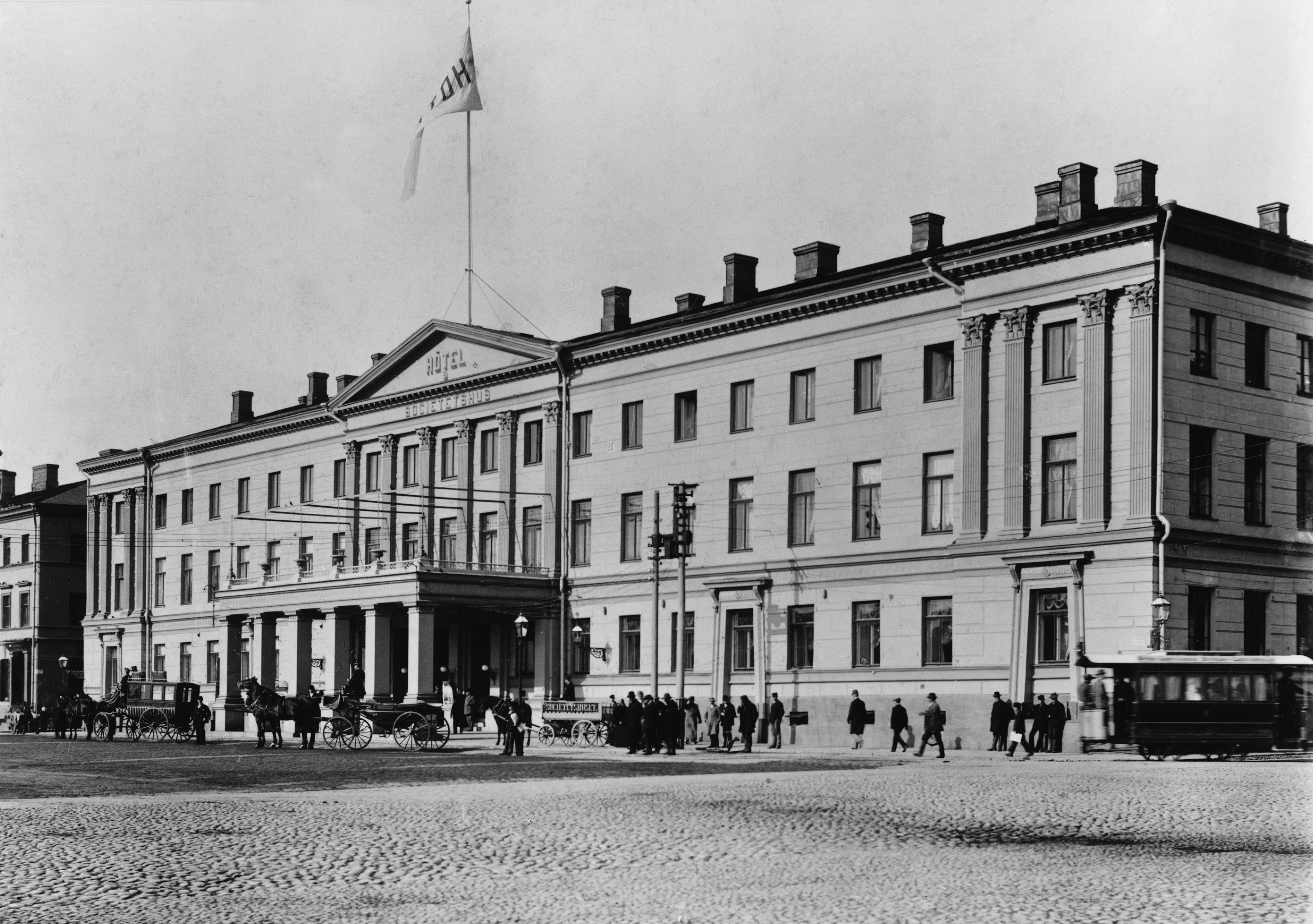 Arkistokuva: Mustavalkoinen valokuva Helsingin kaupungintalon julkisivusta, jossa on pylväitä ja parveke, sekä kadusta hevoskärryineen ja ihmisineen.