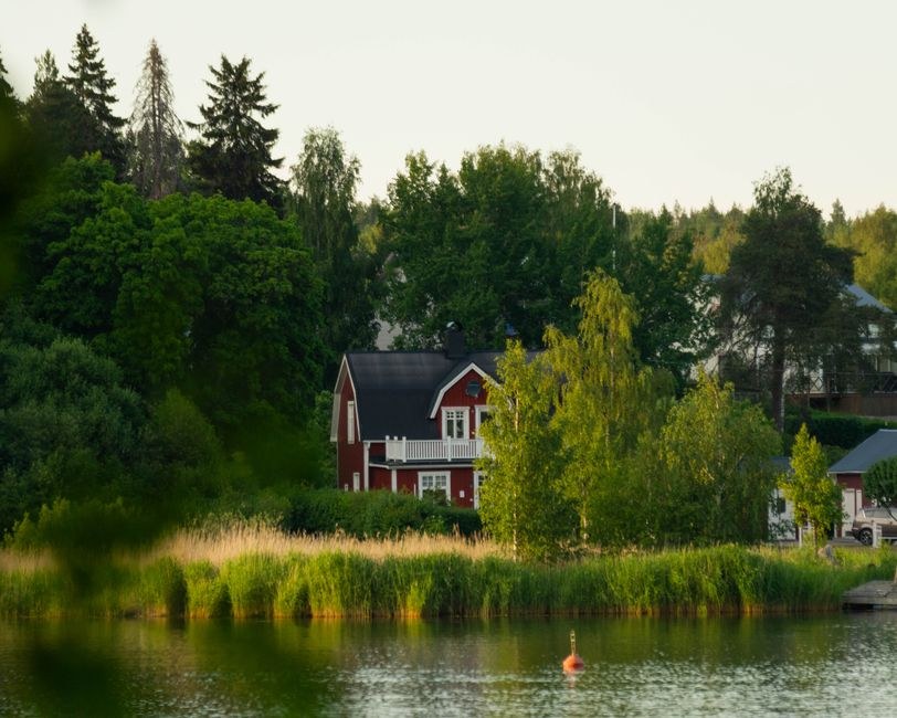 Arkistokuva: Punainen puumökki valkoisilla yksityiskohdilla ja tummalla katolla seisoo järven rannalla vihreiden puiden ja korkeiden ruokojen ympäröimänä.