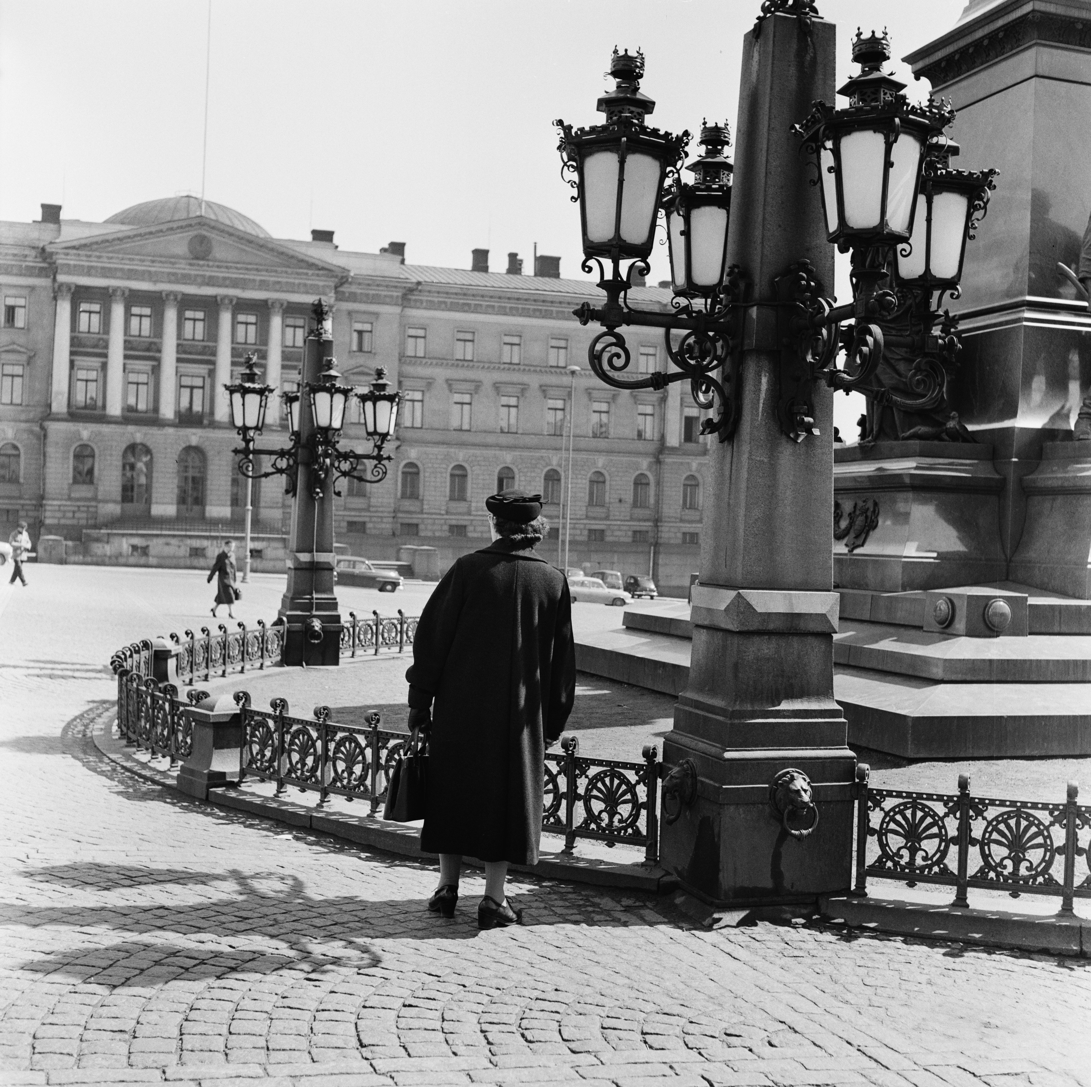 Arkistokuva: Nainen kävelee selin kohti suurta rakennusta, jonka edessä on katuvalaisimia ja koristeellinen metalliaita.
