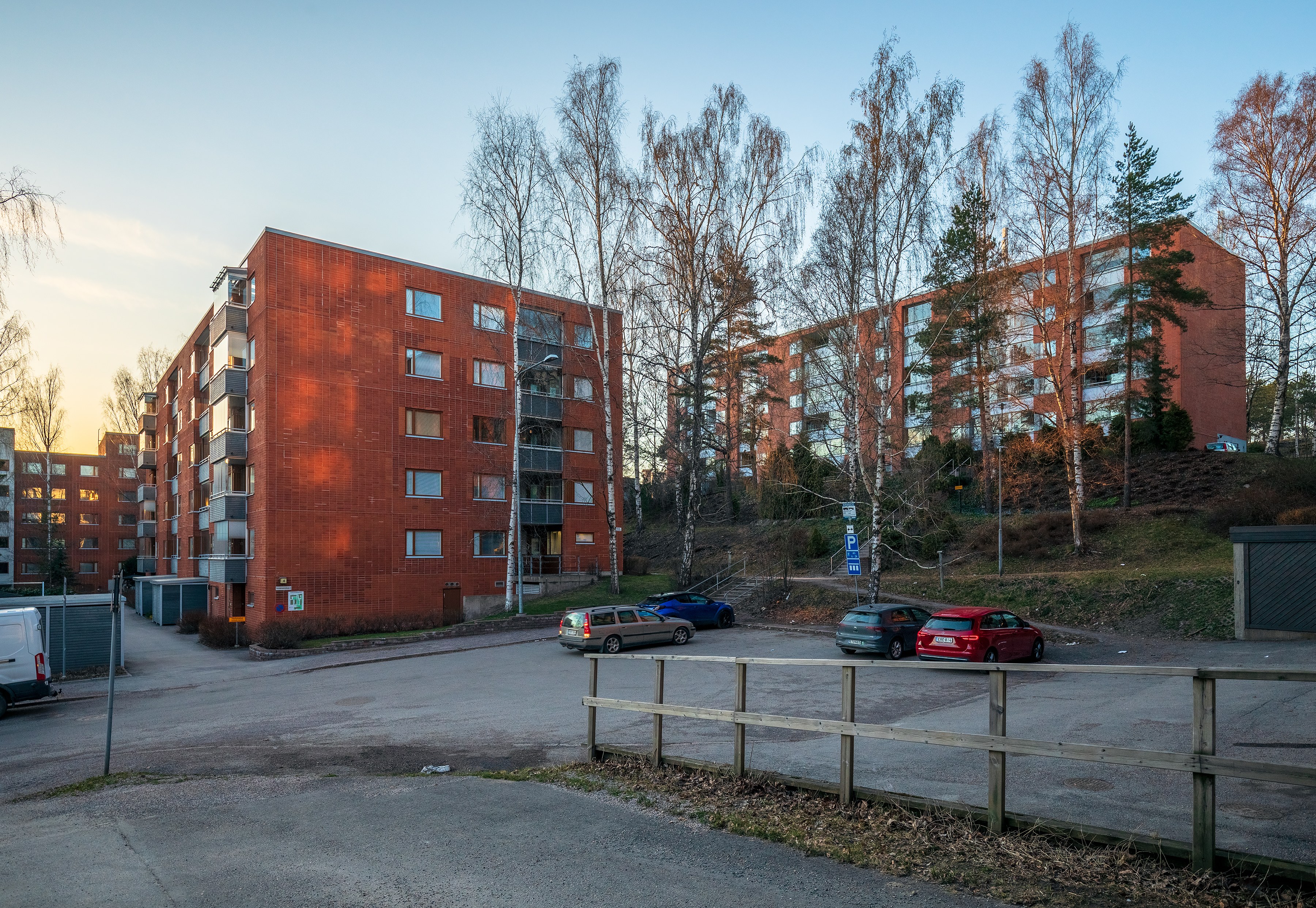 Arkistokuva: Useita punatiilisiä kerrostaloja ja paljaita puita asuinalueen kadun ja pysäköintialueen ympärillä ilta-auringossa.