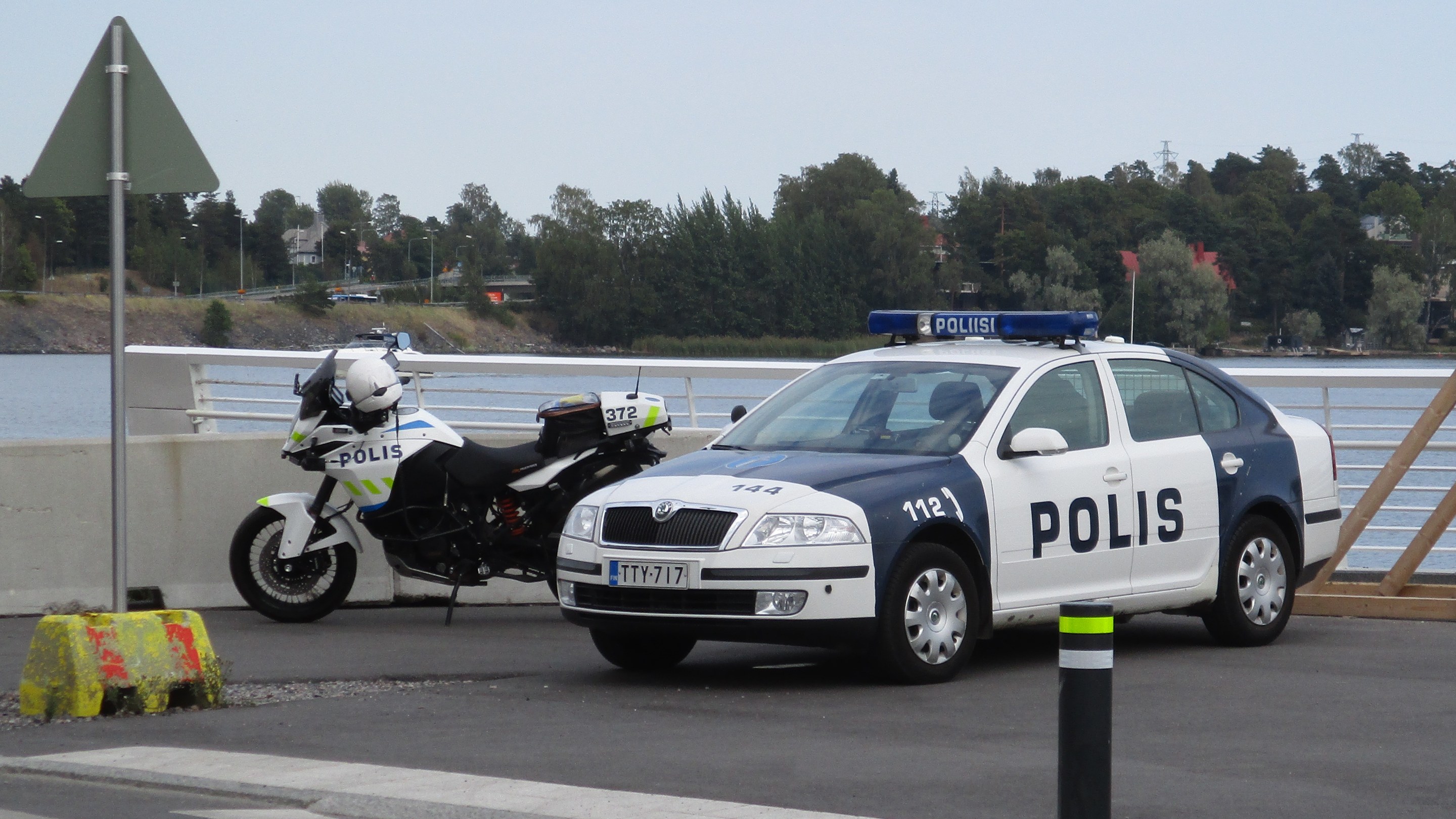 Arkistokuva: Valkoinen ja tummansininen poliisiauto sekä valkoinen poliisimoottoripyörä ovat pysäköityinä asfalttipihalla veden äärellä.
