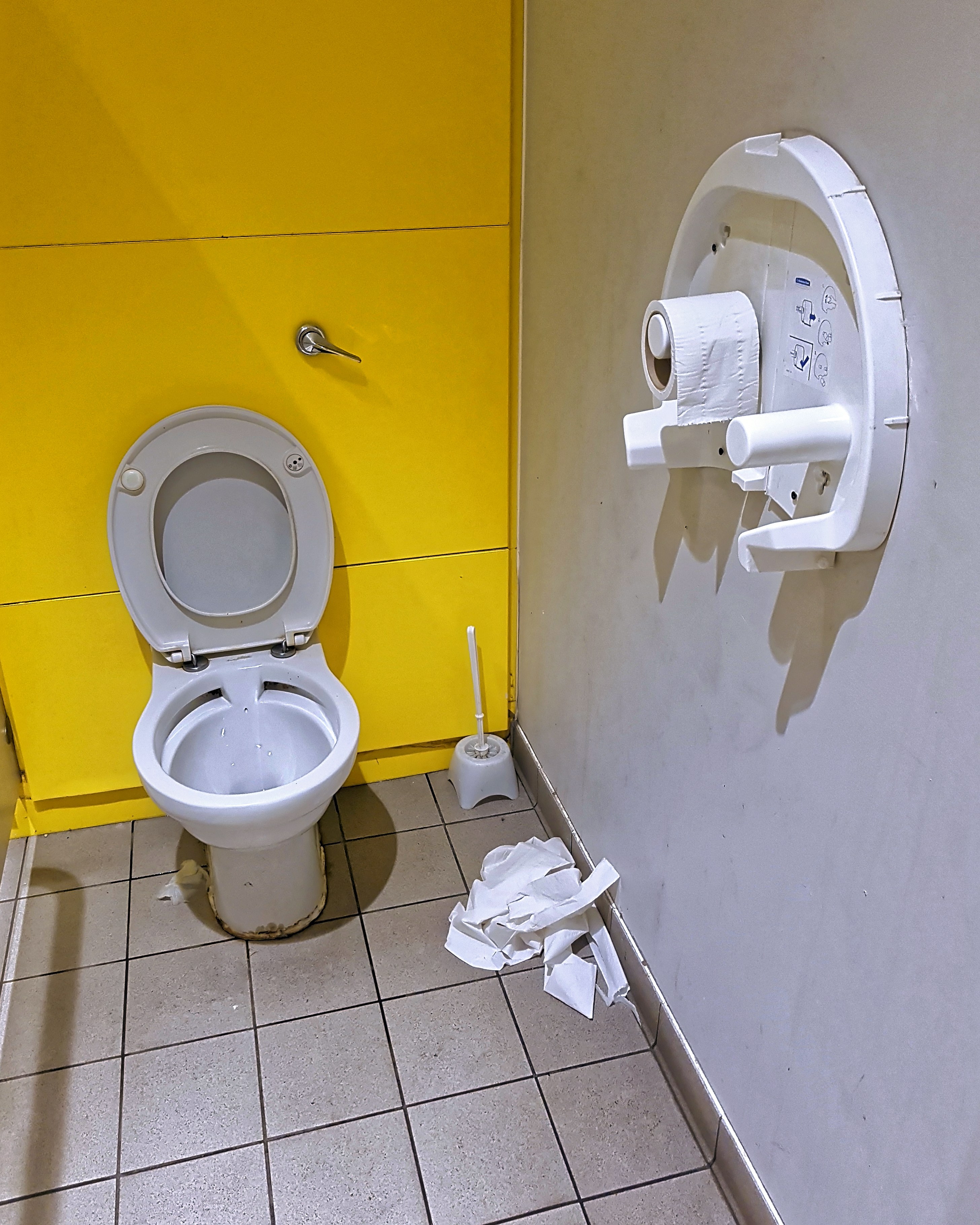 Arkistokuva: Yleisen wc-tilan sisällä on keltainen seinä, valkoinen wc-istuin ja harmaat laattalattiat. Seinällä on wc-paperiteline.