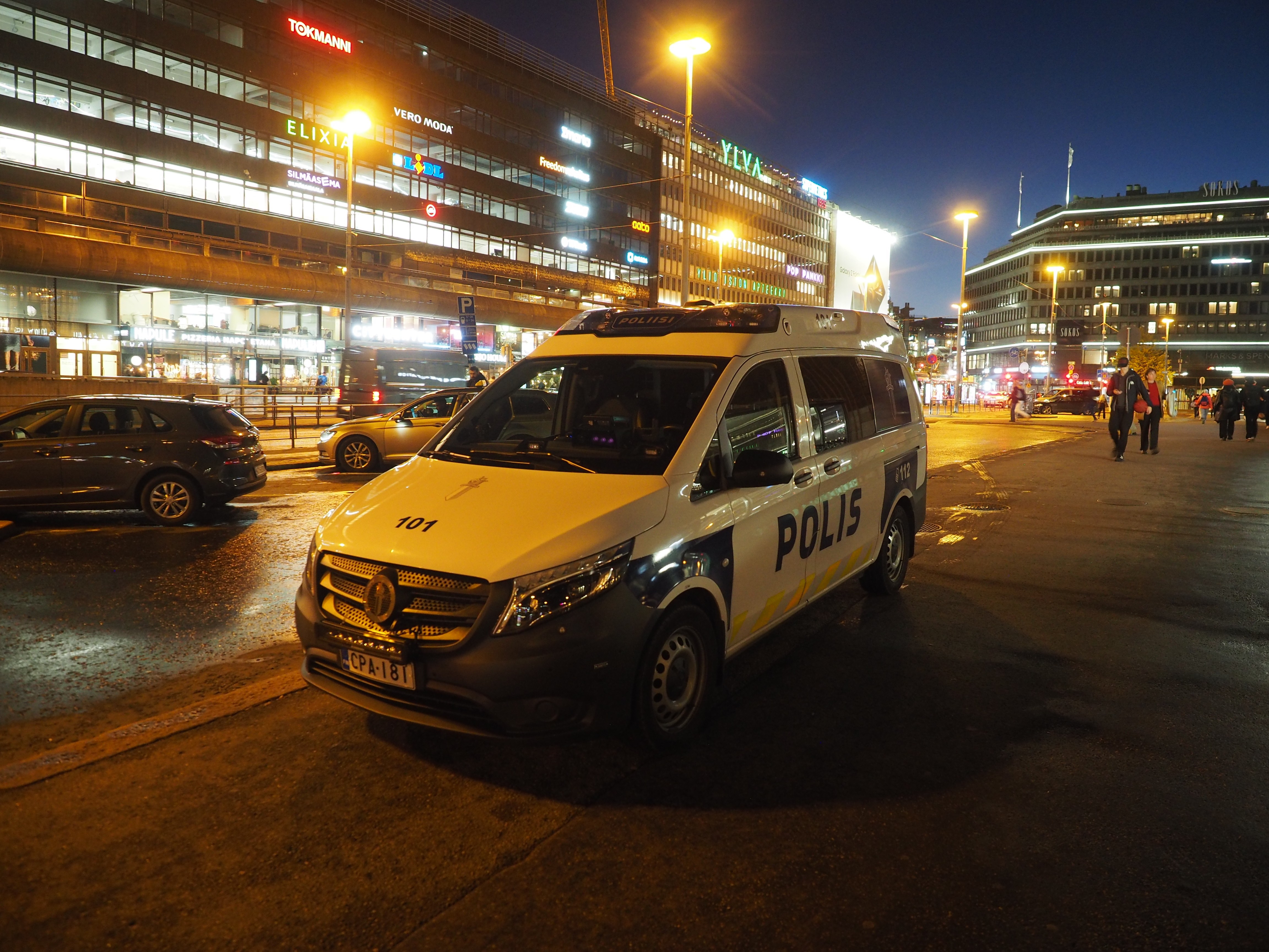 Arkistokuva: Valkoinen ja sininen poliisin partioauto on pysäköitynä kaupungin kadulle iltavalaistuksessa, taustalla näkyy valaistuja rakennuksia ja katuvaloja.