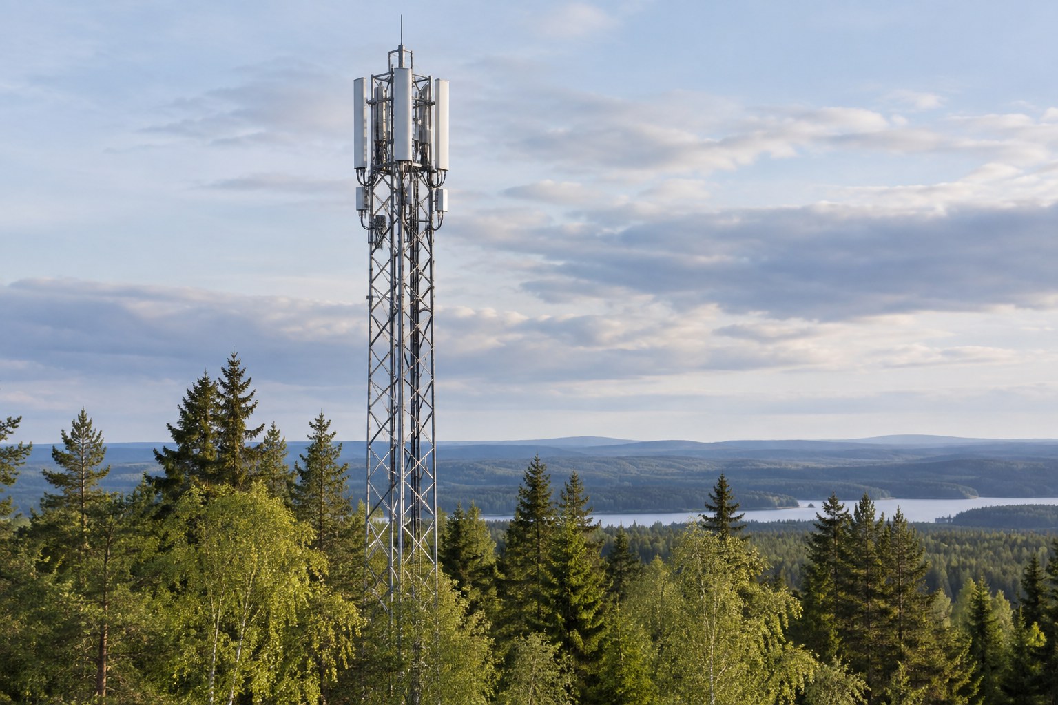 AI-generoitu kuva: Teräksinen matkaviestinverkon tukiasemamasto kohoaa metsäisessä maisemassa, jossa näkyy myös järvi ja kaukaisia kukkuloita.