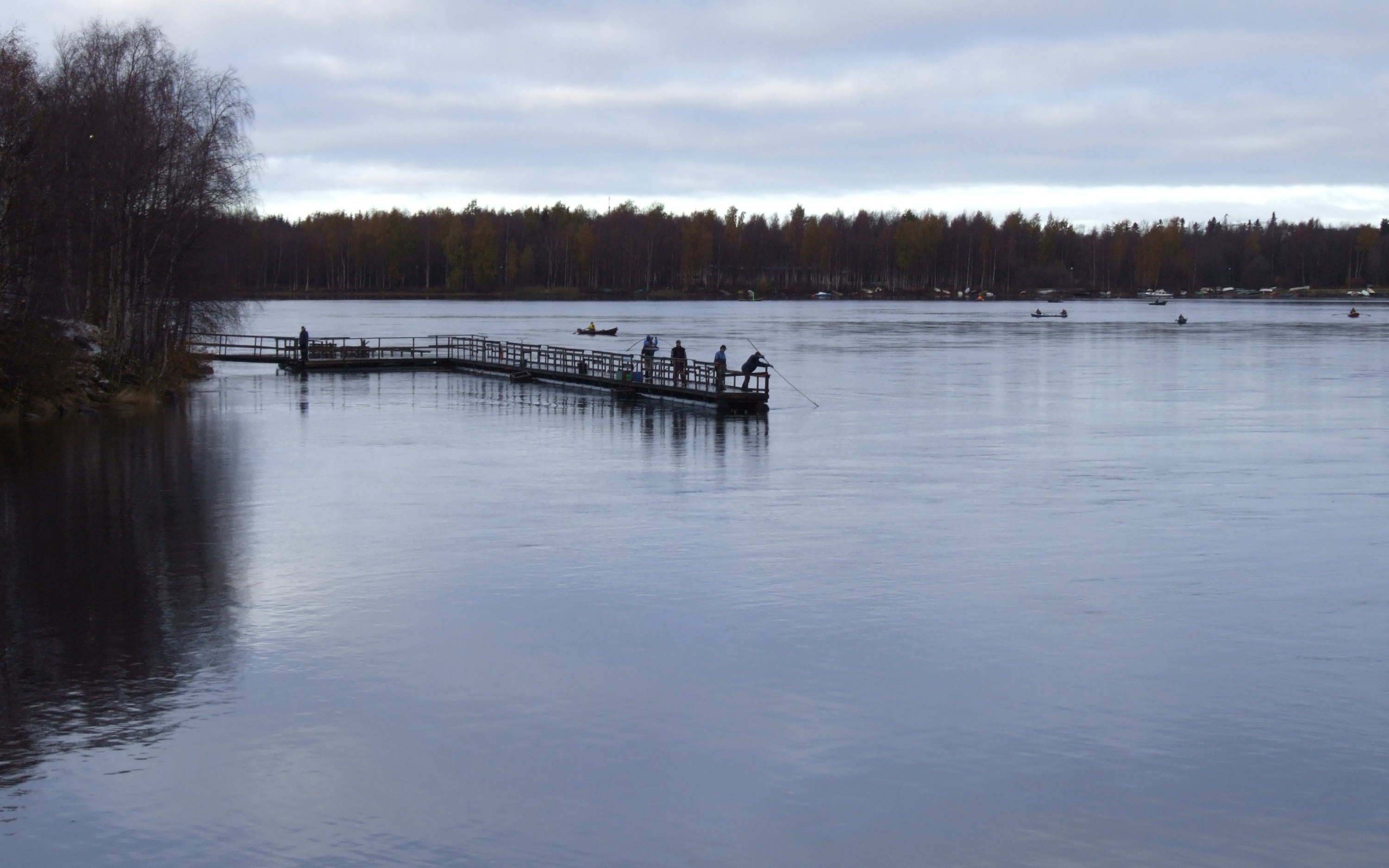 Arkistokuva: Laaja näkymä joesta, jossa on pitkä puinen laituri ja useita ihmisiä. Yksi henkilö käyttää pitkävartista lippoa vedessä.
