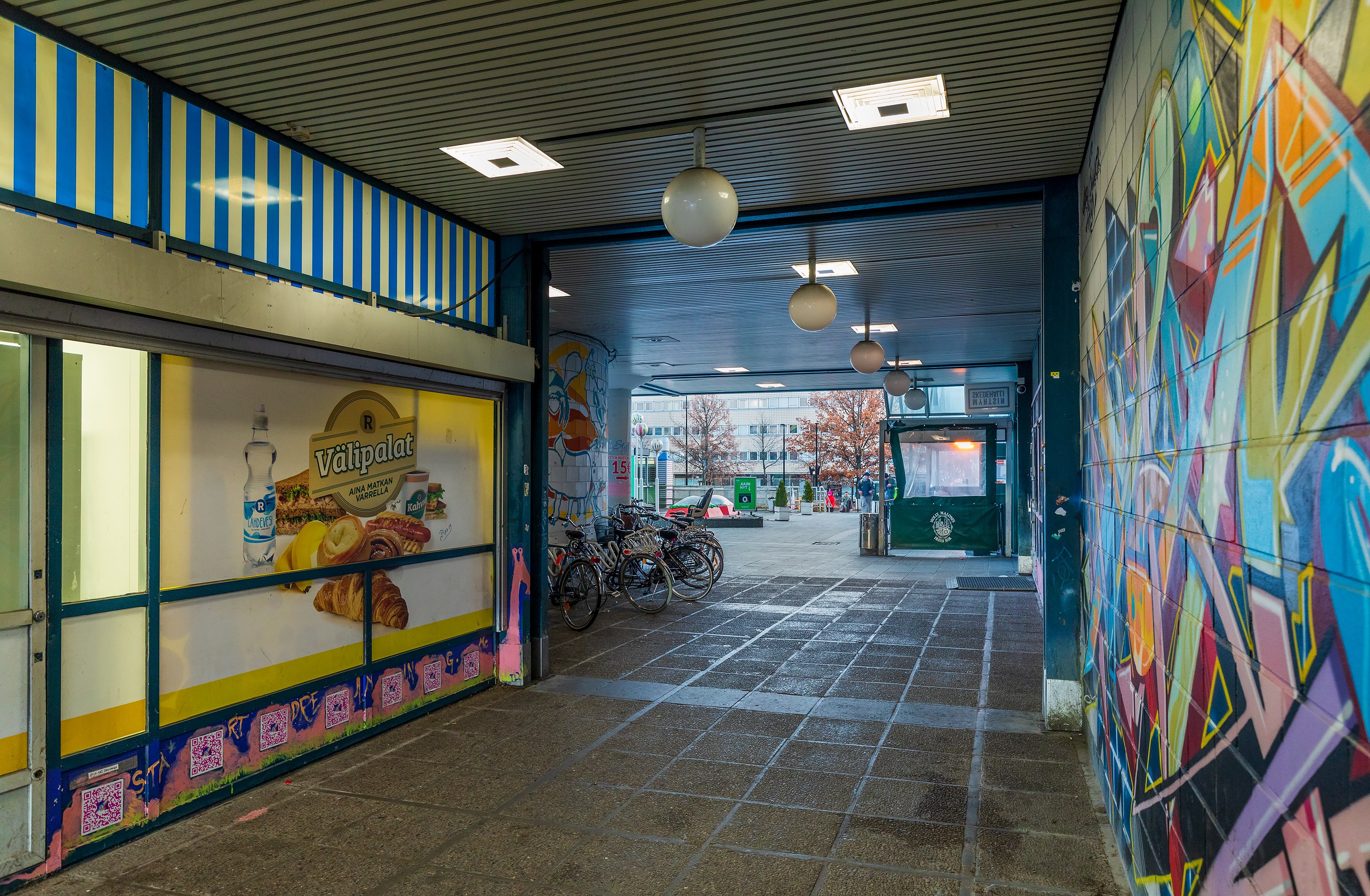 Arkistokuva: Käytävä, jossa on laattalattia ja katettu yläosa, johtaa ulkoalueelle. Vasemmalla on mainoksia sisältävä liiketila ja oikealla graffitiseinä.